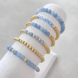 May include: A set of six delicate bracelets featuring light blue gemstone beads and gold beads. The bracelets are stacked on a white display stand.