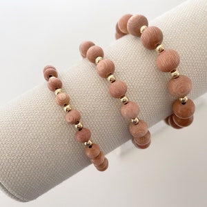 May include: Three wooden bead bracelets with gold accents. The bracelets are stacked on a white fabric display stand.