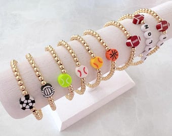 May include: Gold beaded bracelets with sports-themed charms, including a soccer ball, volleyball, tennis ball, baseball, basketball, and football. One bracelet spells out a name with white letter beads. The bracelets are displayed on a white stand.