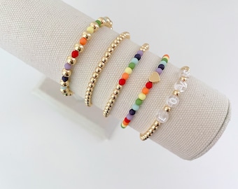 May include: Five gold bead bracelets with colorful beads. One bracelet has a heart charm. One bracelet has clear beads with the word "LOVE" spelled out.