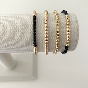May include: Four gold beaded bracelets with black beads on the ends. The bracelets are stacked on a white display stand.