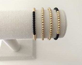 May include: Four gold beaded bracelets with black beads on the ends. The bracelets are stacked on a white display stand.