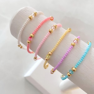 May include: Six colourful beaded bracelets with gold accents. The bracelets are arranged in a row on a white surface. The bracelets are made of small, round beads in shades of pink, yellow, blue, and white.