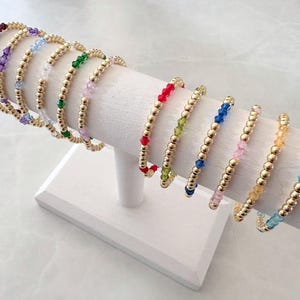 Può includere: Esposizione di braccialetti dorati con accenti di pietre preziose colorate. I braccialetti sono disposti su un espositore bianco, mostrando una varietà di colori tra cui rosso, verde, blu e viola. Ogni braccialetto misura circa 18 cm di circonferenza.