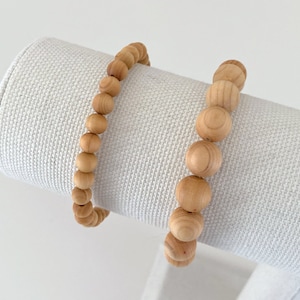 May include: Two wooden bead bracelets on a white fabric display stand. The bracelets are made of natural wood beads and are a light brown color.