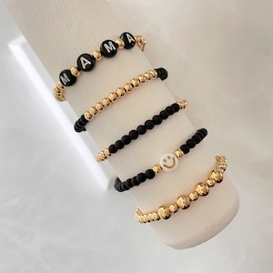 May include: Four bracelets with black and gold beads. One bracelet has the word "MAMA" spelled out in white letters on black beads. Another bracelet has a white smiley face charm. The other two bracelets are solid gold.