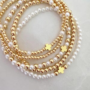 Op de afbeelding: Meerdere gouden armbanden met witte parels en gouden kruisbedels. De armbanden zijn gelaagd, waardoor een gestapelde look ontstaat. De kralen zijn klein en rond, en de kruizen zijn plat en eenvoudig.