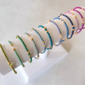 May include: A set of 10 delicate beaded bracelets in various shades of green, blue, and pink. Each bracelet features small gold beads and a single crystal bead.