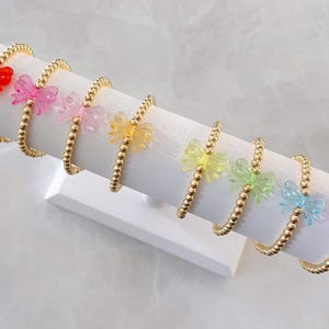 May include: A set of seven gold beaded bracelets with colorful bow charms. The bows are in shades of red, pink, yellow, green, blue, and purple.