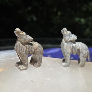 Puede incluir: Dos figuras de lobos talladas en piedra. Los lobos son grises con vetas marrones y beige, en pose de aullido con la cabeza hacia atrás. Los ojos están resaltados con un color dorado. Las figuras están sobre una superficie blanca.