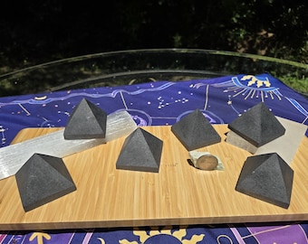 May include: A collection of black pyramid-shaped stones, arranged on a wooden board. A long, white crystal and a small, round stone are also present. The board rests on a blue cloth with a celestial pattern.