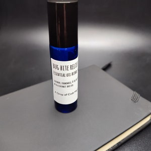 Puede incluir: Un frasco de vidrio azul con tapa negra. La etiqueta dice "Bug Bite Relief Essential Oil Blend, Lavender, Chamomile, & Tea Tree with a Coconut Base Oil, A Drop of Craft Magic."