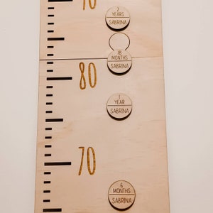 May include: A wooden growth chart with a ruler marked in centimetres and inches. The chart has a light wood finish and features round wooden markers with the text "Sabrina" and the child's age in months or years. The markers are placed at different heights on the chart, indicating the child's growth over time.