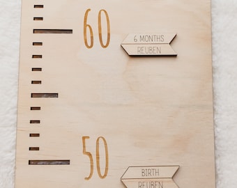 May include: A wooden growth chart with a ruler marked in centimetres. The chart has two wooden arrows with text. The top arrow says "6 months Reuben" and the bottom arrow says "Birth Reuben".