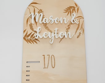 May include: A wooden growth chart with a light brown background and a white arch at the top. The arch is decorated with a floral design and the names "Mason & Leyton" are written in white letters. The chart has a height measurement scale with the number "170" marked on it.