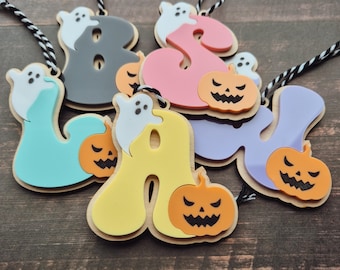 May include: A collection of colorful Halloween-themed letter ornaments. Each letter is a different color, with a ghost and jack-o'-lantern design. The letters are attached to a black and white string for hanging.