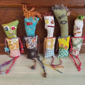 May include: Five handmade fabric dolls with whimsical faces and button eyes. Each doll has a unique color scheme, with patterned fabric bodies and yarn or fabric hair. The dolls are arranged on a wooden surface.