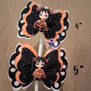 May include: Two decorative butterfly hair bows. The bows are black with orange glitter accents and white edges. Each bow features a small doll in an orange dress. One bow is 4 inches, the other is 5 inches.