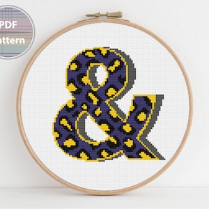 Può includere: Schema di punto croce del simbolo ampersand in un design con stampa leopardata viola e gialla. Lo schema è mostrato in un telaio da ricamo in legno.