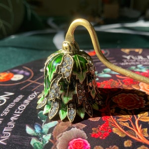 May include: A gold candle snuffer with a green and white enamel bell-shaped top decorated with rhinestones. The bell is decorated with a leaf pattern. The snuffer is sitting on a black and red floral patterned surface.