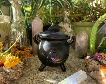 May include: A small, black cast iron cauldron with a lid, three legs, and two handles. The cauldron features a decorative design on the front. The background includes crystals, plants, and dried flowers, creating a mystical aesthetic.