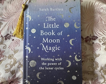 May include: A hardcover book titled "The Little Book of Moon Magic" by Sarah Bartlett. The cover features a gradient of blue and purple with silver crescent moons and stars. A gold tassel bookmark is attached.