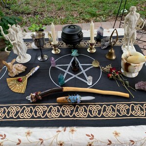 May include: A black altar cloth with a pentagram design and gold trim. There are two white statues, a cauldron, candles, crystals, a goblet, a wand, a knife, and a broom on the cloth.