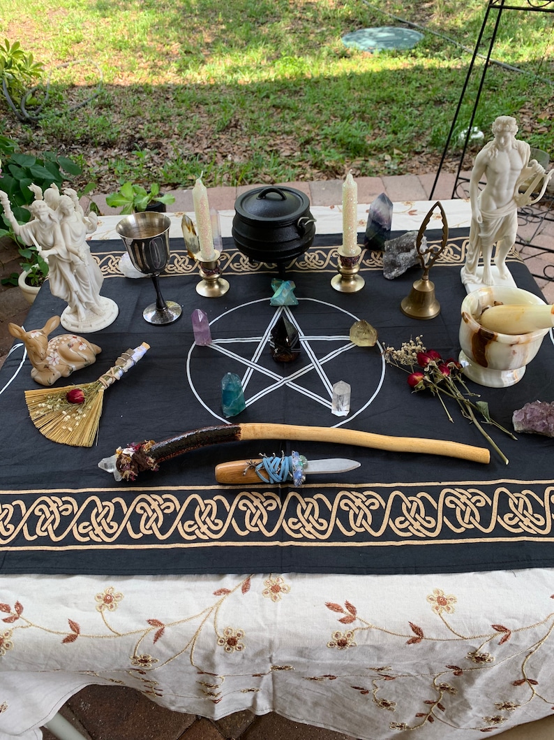 May include: A black altar cloth with a pentagram design and gold trim. There are two white statues, a cauldron, candles, crystals, a goblet, a wand, a knife, and a broom on the cloth.