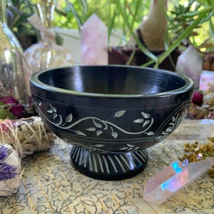 May include: A black slate bowl with a carved floral design. The bowl is on a pedestal and has a smooth, polished finish.