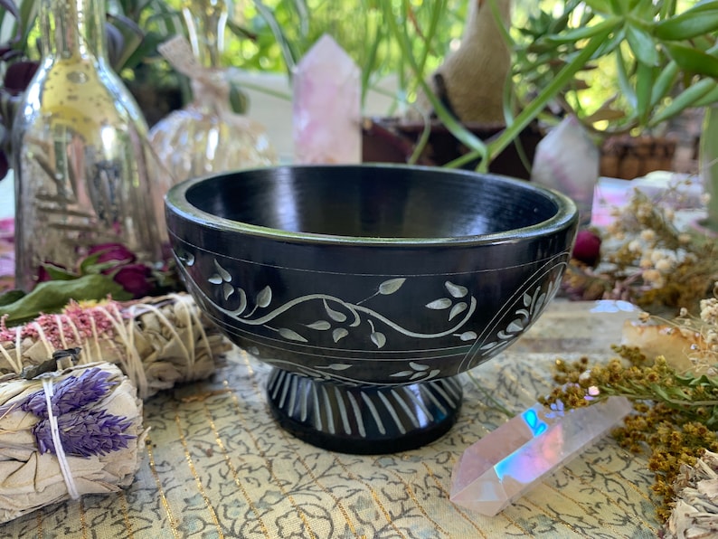 May include: A black slate bowl with a carved floral design. The bowl is on a pedestal and has a smooth, polished finish.
