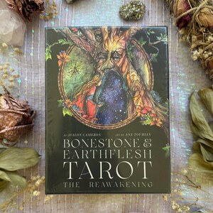May include: A tarot card deck titled "Bonestone & Earthflesh Tarot: The Reawakening" with a detailed illustration of a tree-like face on the cover. The deck is surrounded by dried flowers, crystals, and other decorative elements.