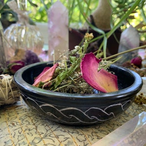 May include: A black slate bowl with a carved design sits on a patterned surface. The bowl is filled with dried rose petals and herbs.