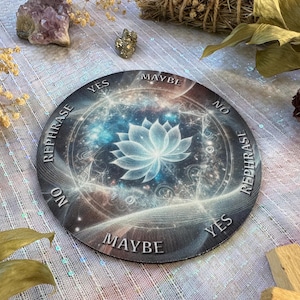 May include: A round divination board with a celestial design featuring a lotus flower in the center. The board has text that reads "Yes", "No", "Maybe", and "Rephrase". The color scheme includes blues, whites, and grays.