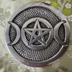 May include: A silver-colored, circular decorative piece featuring a central pentagram within a circle, flanked by crescent moon shapes. The background has a textured, dotted pattern. The piece is likely used for spiritual or decorative purposes.