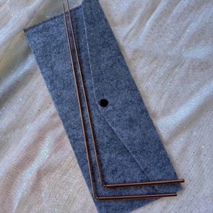 May include: A gray felt case with a black button closure holds two copper-colored metal rods. The rods are straight and bent at a 90-degree angle. The case and rods rest on a shimmering, light-colored fabric.