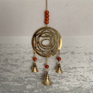 May include: A brass-colored wind chime with a circular design and three small bells. The chime features a series of orange beads and a gold-colored chain for hanging. The design has an intricate pattern.