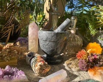 May include: A gray marble mortar and pestle set on a table with various crystals, dried flowers, and a smudge stick.