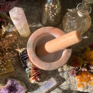 May include: A pink and gray mortar and pestle set with a white crystal next to it. The mortar and pestle are made of natural stone and have a rough texture. The crystal is clear and has a smooth surface. The set is surrounded by dried flowers and other crystals.
