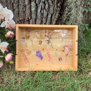 May include: A wooden tray with a clear resin top featuring pressed flowers in shades of pink, purple, and brown. The tray has black metal handles.
