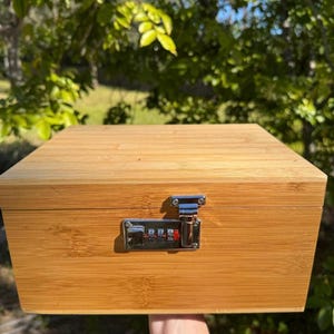 May include: A wooden box with a combination lock. The box is made of light brown wood and has a natural finish. The lock is silver and has a combination dial.