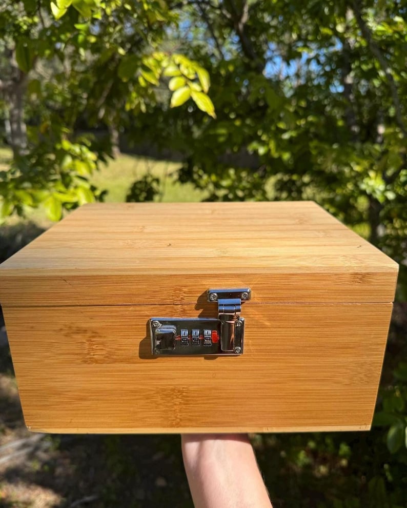 May include: A wooden box with a combination lock. The box is made of light brown wood and has a natural finish. The lock is silver and has a combination dial.