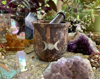 Puede incluir: Un mortero y una maja marrones y beiges con un diseño de triple luna. El mortero está rodeado de cristales, plantas y flores secas. La maja descansa dentro del mortero. El mortero y la maja probablemente se utilizan para moler hierbas y otros ingredientes.