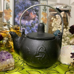 May include: A small black cast iron teapot with a silver handle and a Celtic knot design. The teapot is sitting on a green and gold patterned surface.