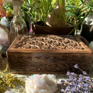 May include: A rectangular wooden box with intricate floral carvings on the lid. The box is a warm brown color and appears to be made of wood. The box is surrounded by other decorative items.