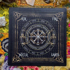 May include: A black divination board with gold-colored accents. The board features a circular design with astrological symbols, days of the week, and the words "Yes," "No," and "Maybe." The corners have decorative Celtic knot designs. The words "Air," "Fire," "Water," and "Earth" are also visible.