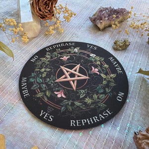 May include: A black round divination board with a pentagram in the center, surrounded by floral and leaf designs. The words "Yes", "No", "Maybe", and "Rephrase" are printed around the edge. The board is decorated with pink flowers and green leaves.