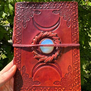 May include: A red leather journal with a triple moon design and a moonstone inlay. The journal has a decorative border and a leather closure.