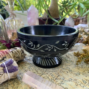 May include: A black soapstone bowl with a carved floral design. The bowl has a wide, round base and a slightly flared rim. It is perfect for holding incense, crystals, or other small items.