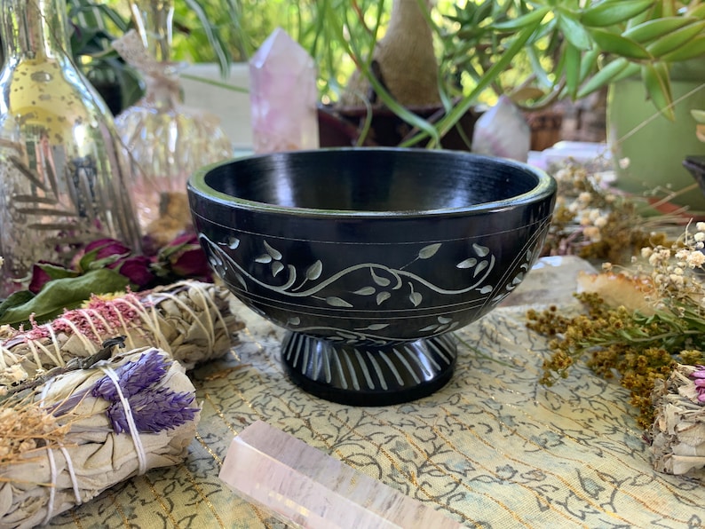 May include: A black soapstone bowl with a carved floral design. The bowl has a wide, round base and a slightly flared rim. It is perfect for holding incense, crystals, or other small items.