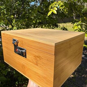 May include: A wooden box with a combination lock. The box is made of light brown wood and has a natural finish. The lock is silver and has a combination dial.